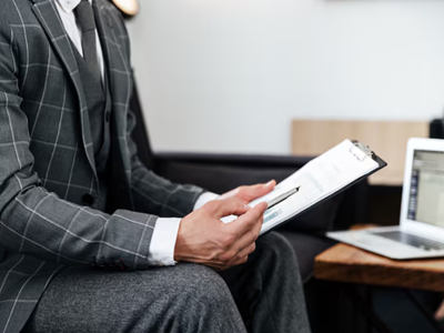 Close-up of a person wearing a grey plaid three-piece suit holding a pen and reviewing papers on a clipboard next to a laptop.